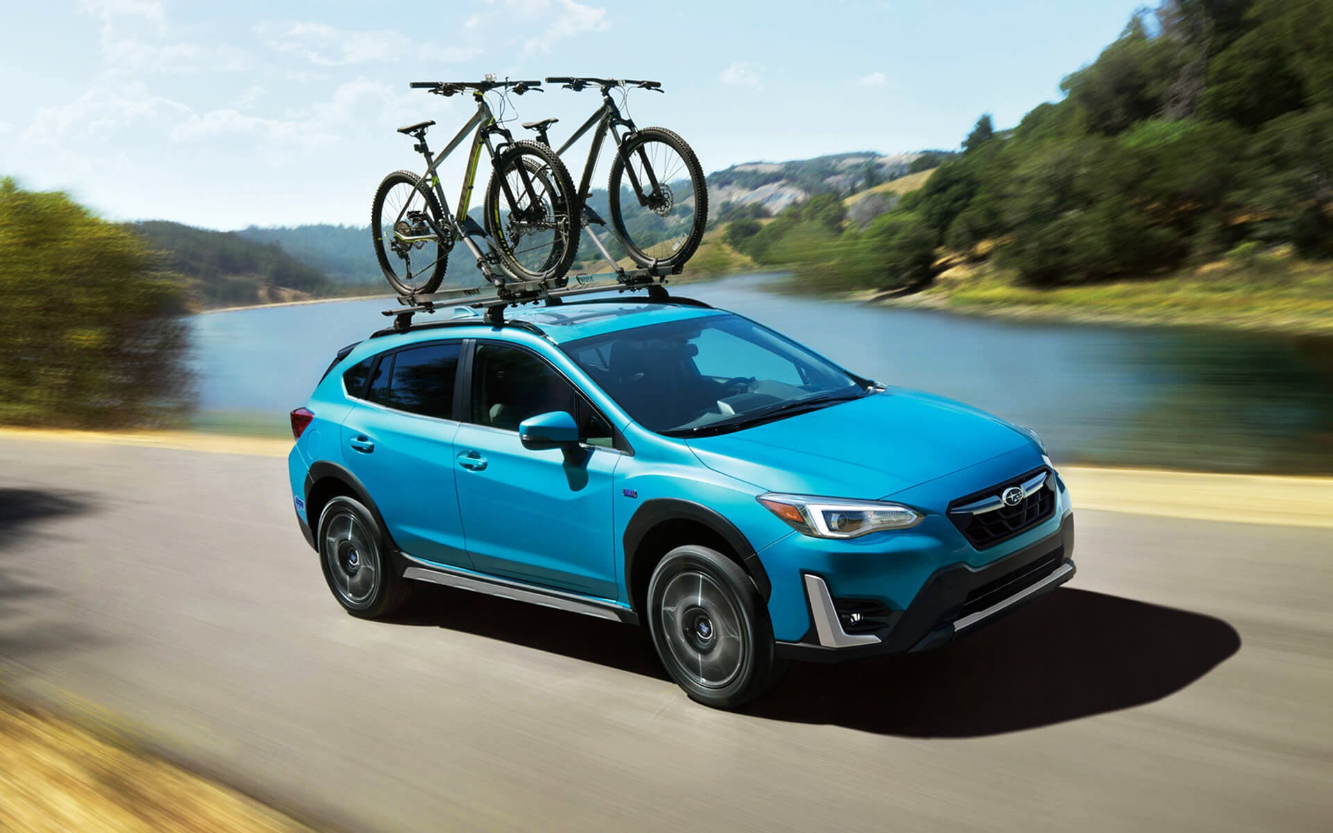 A blue Crosstrek Hybrid with two bicycles on its roof rack driving beside a river | Puente Hills Subaru in City of Industry CA